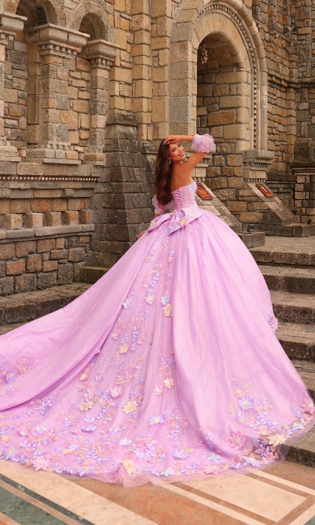 Wearing the Amarra 54234 Puff-Sleeve Quinceanera Dress, a woman in an ornate lavender ball gown with floral lace stands on stone steps before a historic building, glancing over her shoulder.